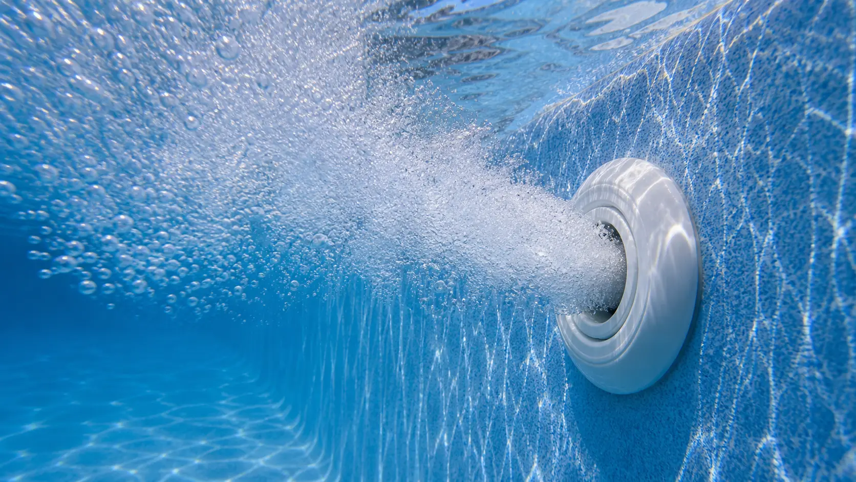 A pool return jet pushes a stream of white air bubbles into clear blue water against a plaster wall.