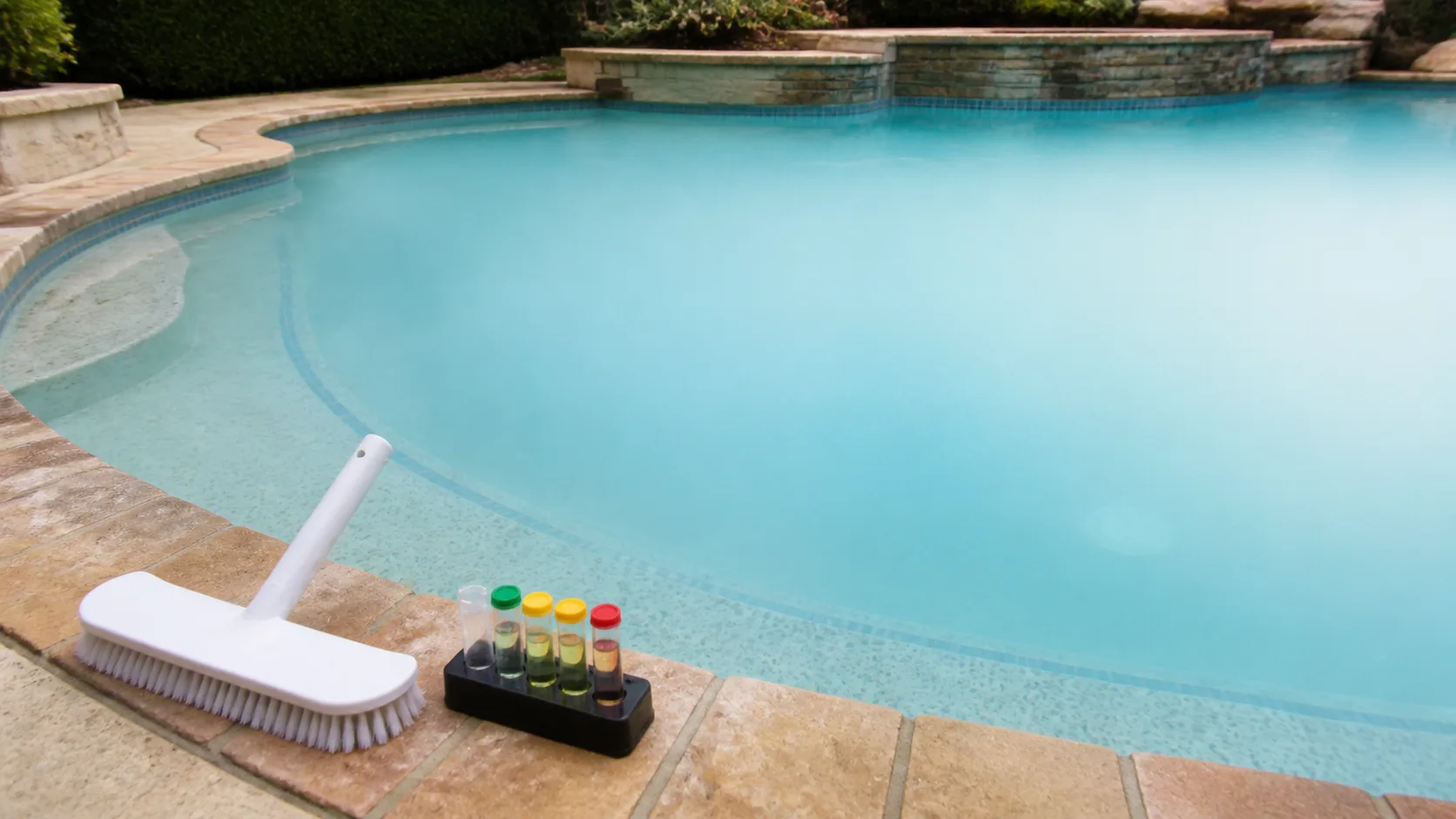 Milky blue pool water obscures the deep end while a pool brush and water test kit rest on the coping.
