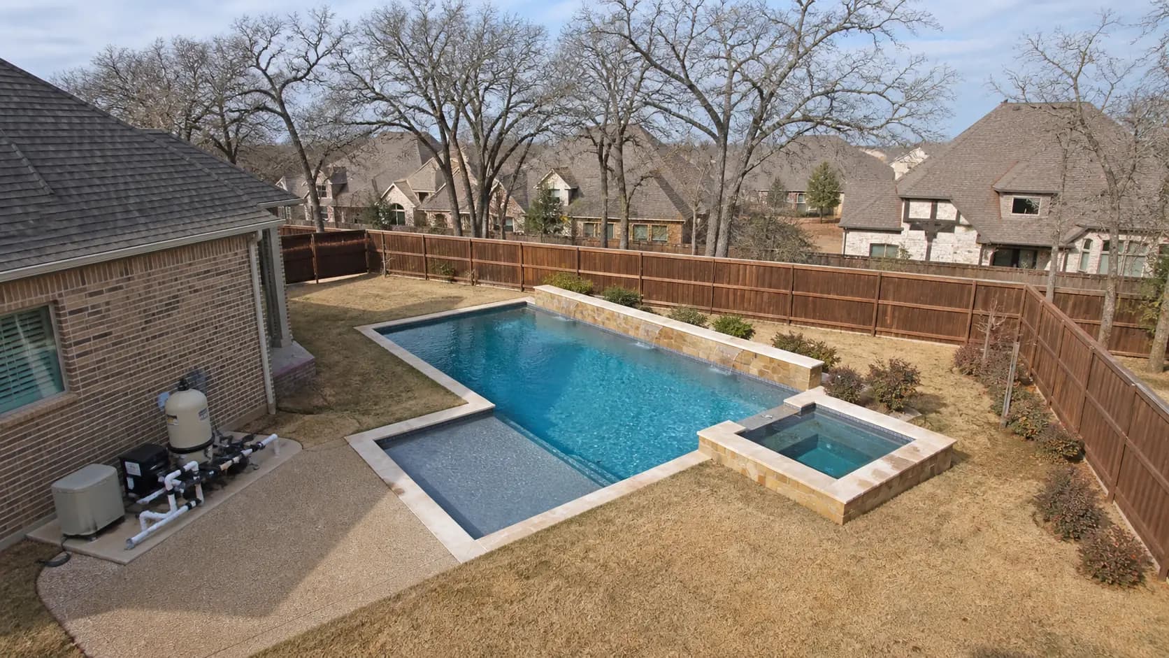 An elevated view over a suburban Collin County backyard with a rectangular pool, small spa, equipment pad against the house, and mature oaks along the fence line.