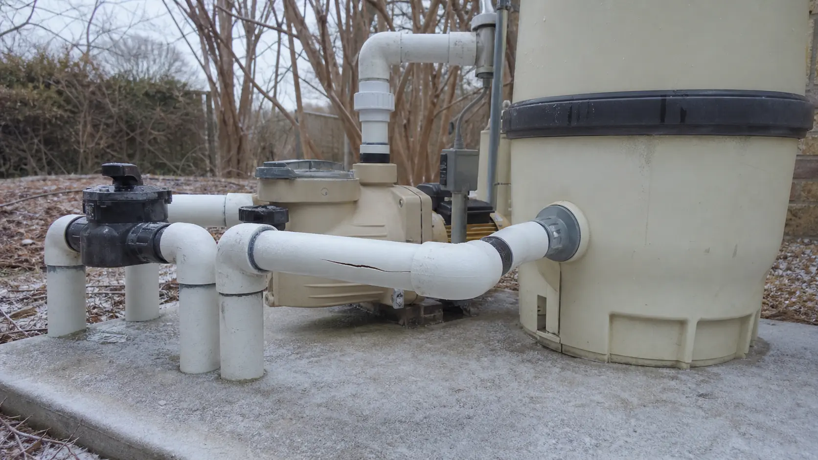 A split running along a white PVC pipe at a frost-covered pool equipment pad, with bare branches behind under overcast winter light.