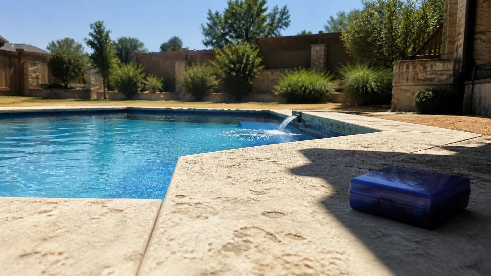 A clear backyard pool circulating under harsh August sun, with heat shimmer over pale stone coping and drought-stressed grass beyond.