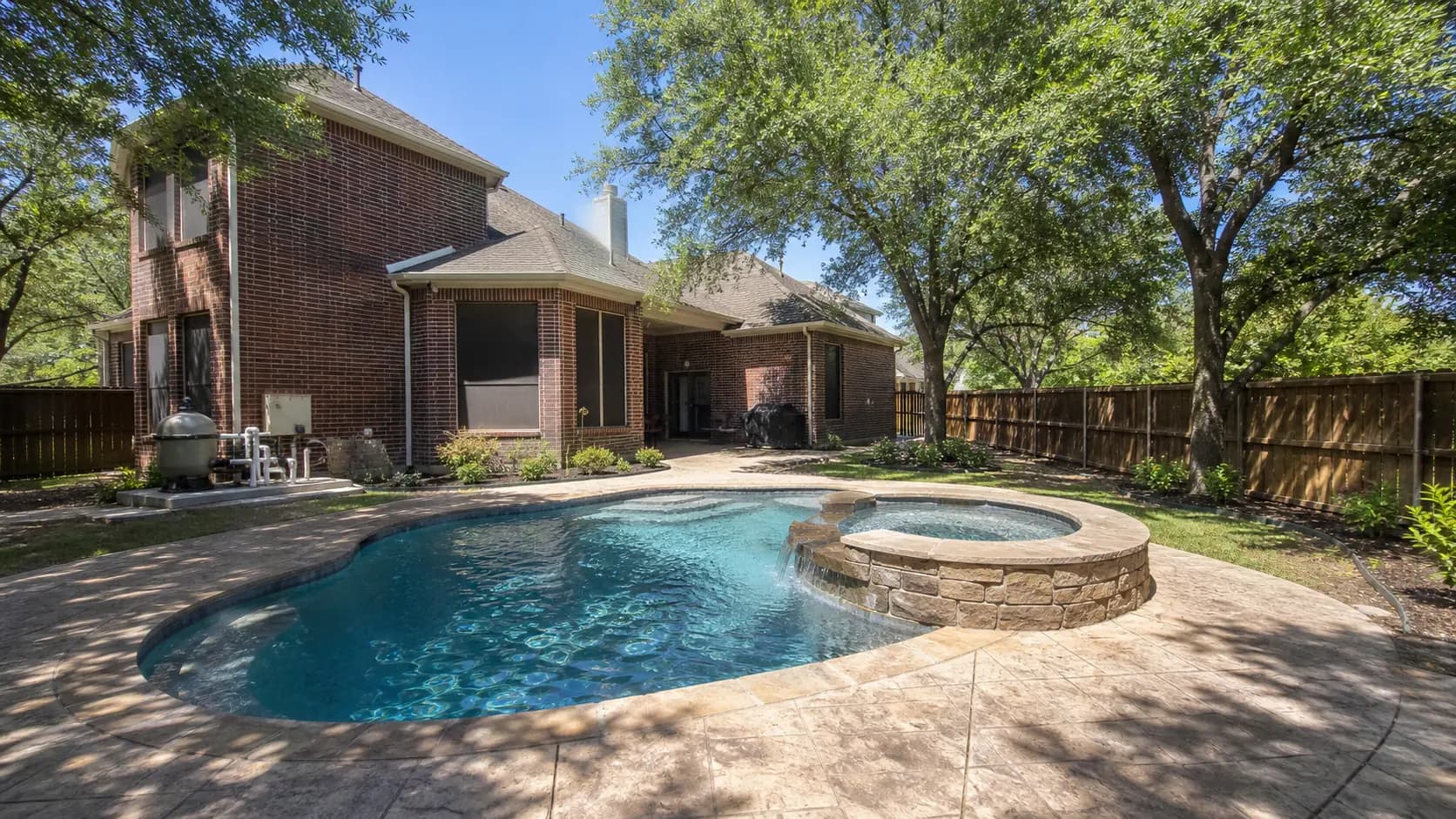 A curved freeform pool with a spillover spa and stamped concrete decking behind a two-story brick home in Allen, shaded by semi-mature live oaks.