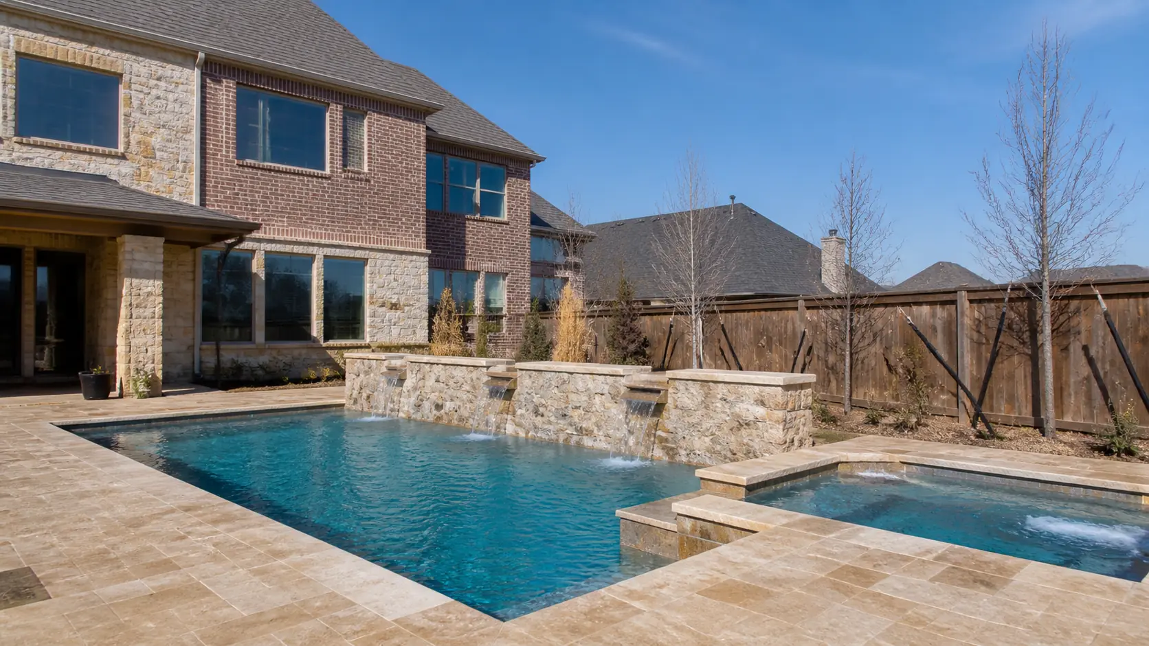 A newer-build rectangular pool with a raised stone water feature wall, travertine decking, and young staked trees behind a large brick and stone Frisco home.