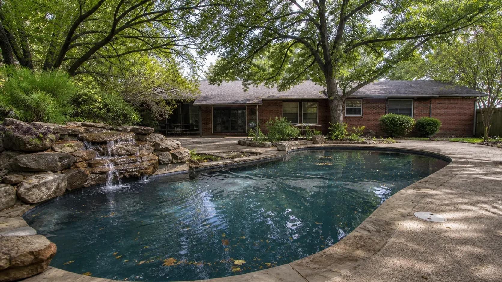 A freeform pool with a rock waterfall behind a red brick ranch home in an established McKinney neighborhood, shaded by mature post oak and pecan canopy.