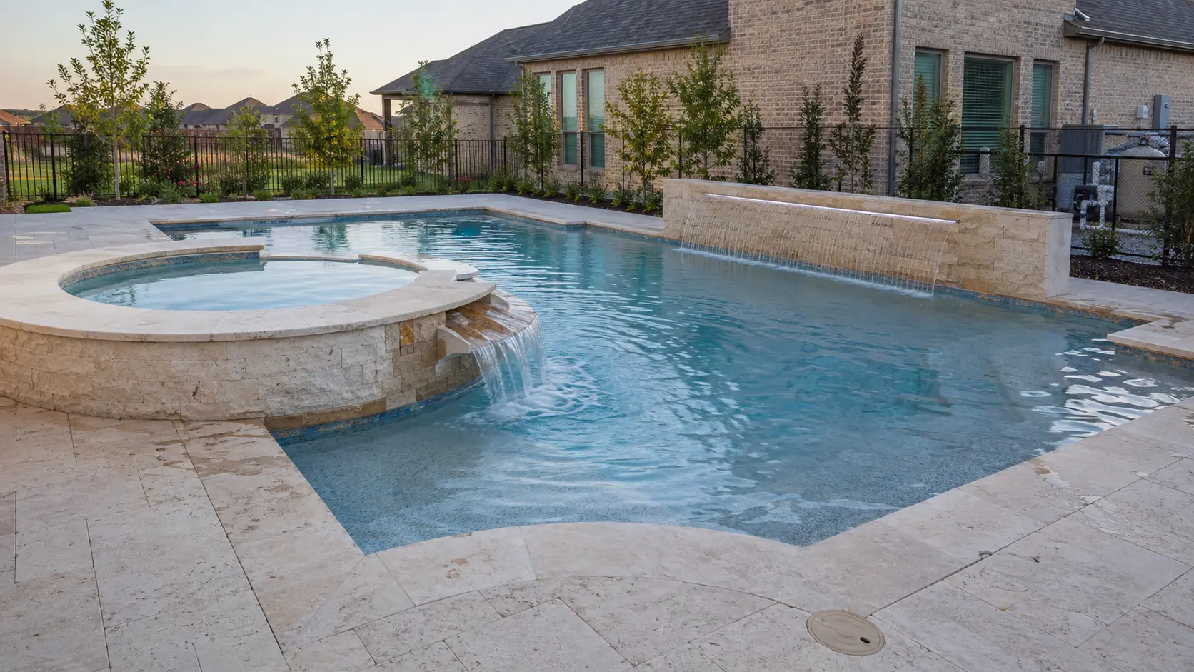 A large Prosper backyard pool at dusk with a raised stone spa spilling into it and a sheer descent running along the far wall, travertine deck and young landscaping around a new brick house.