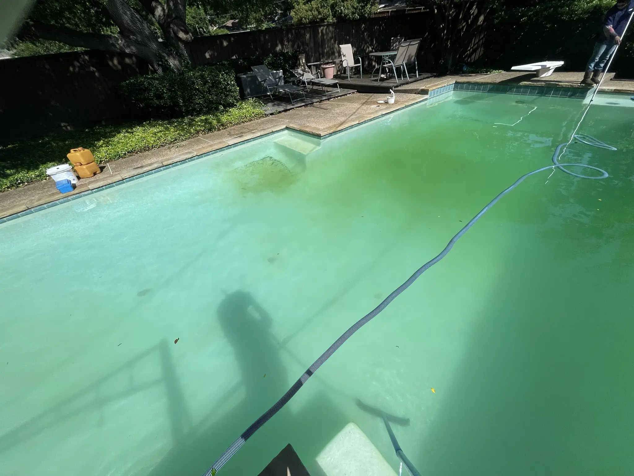 A technician vacuuming a cloudy green rectangular pool with a diving board and chemical containers on the deck.