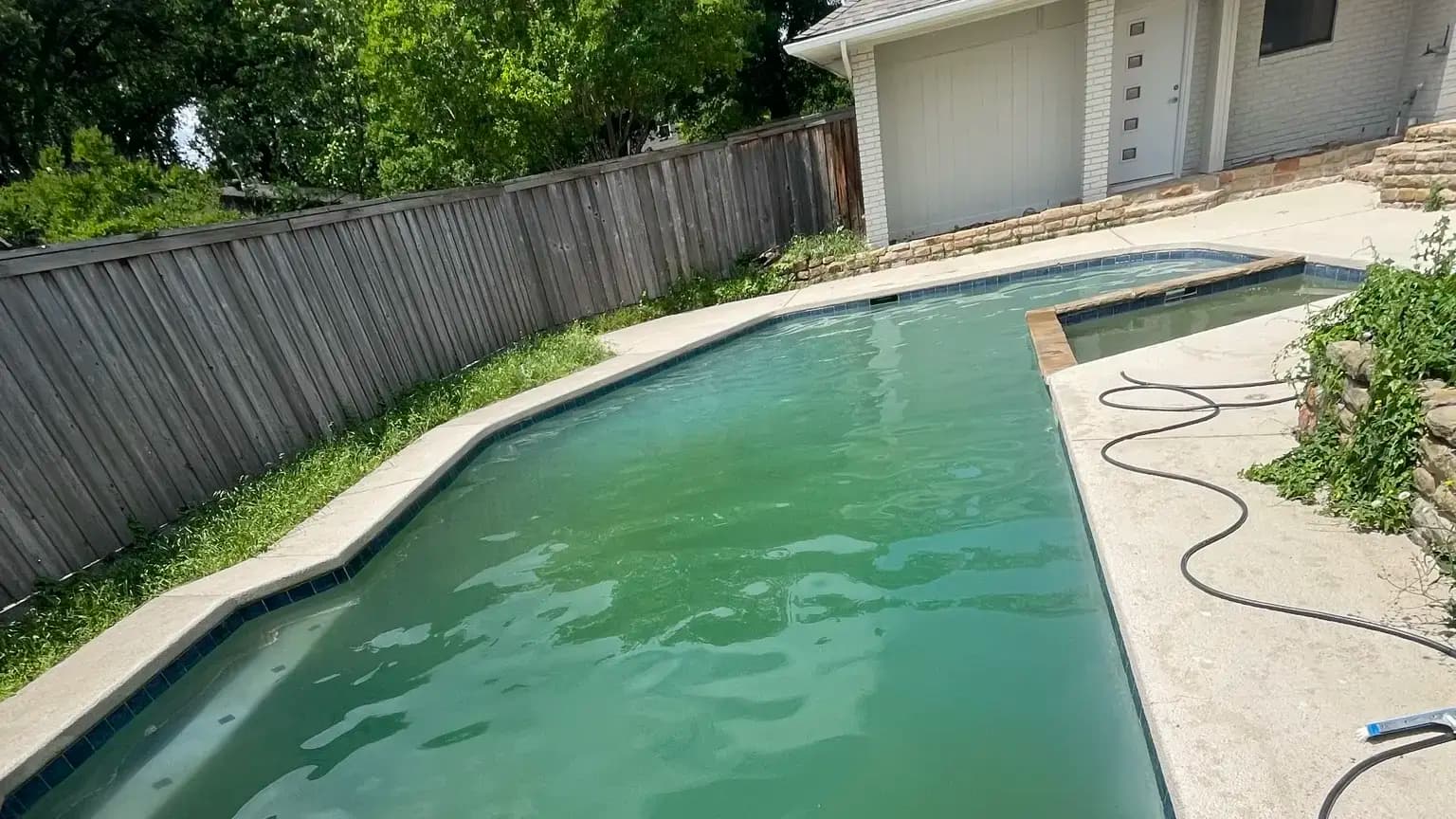 A murky green backyard pool with a raised spa and a garden hose coiled on the deck beside it.