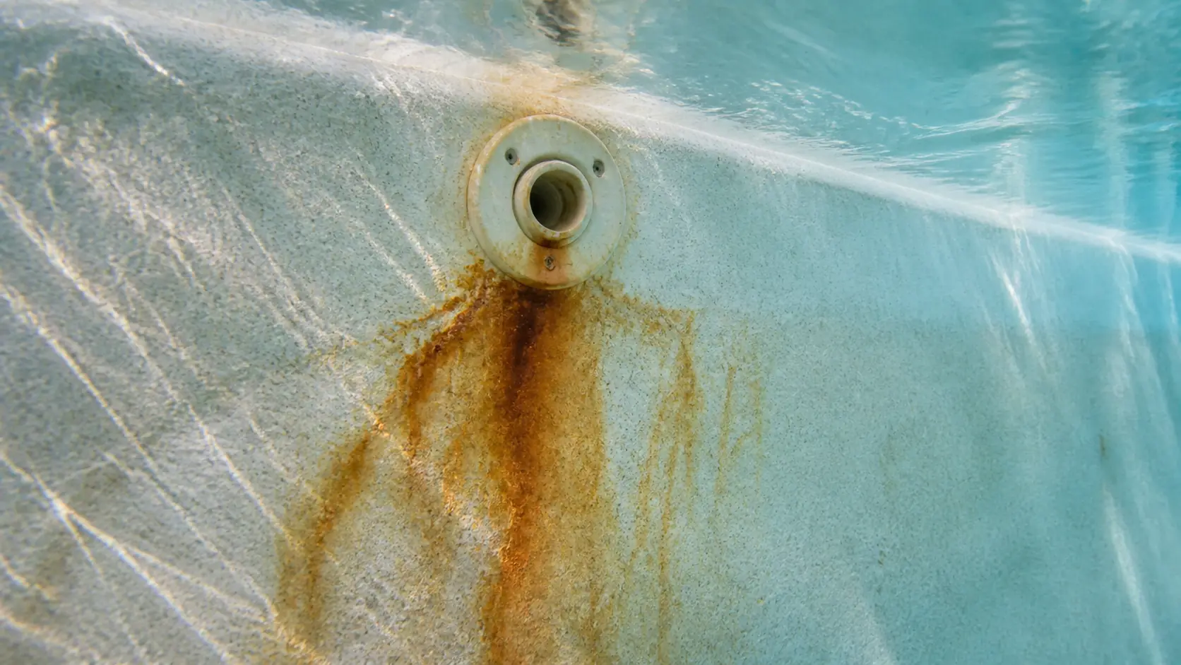 Rust brown metal staining streaking down the pale plaster wall of a swimming pool below a return fitting, seen through clear water.