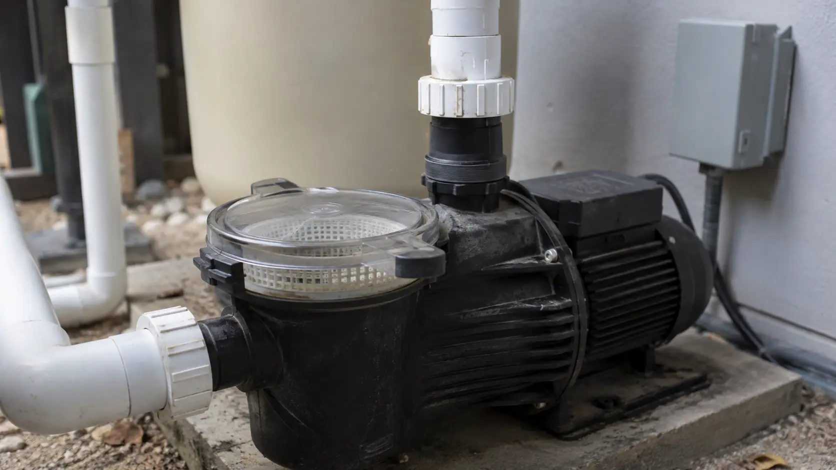 A pool pump and motor on a concrete equipment pad beside a filter tank, with the clear pump lid showing the basket and plumbing unions either side.