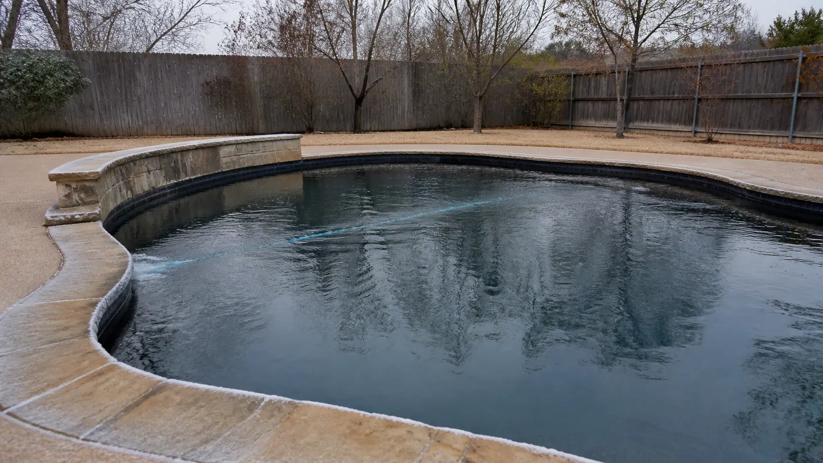 A return jet ripples the surface of a dark winter pool with frost on the shaded stone coping and dormant brown grass beyond.