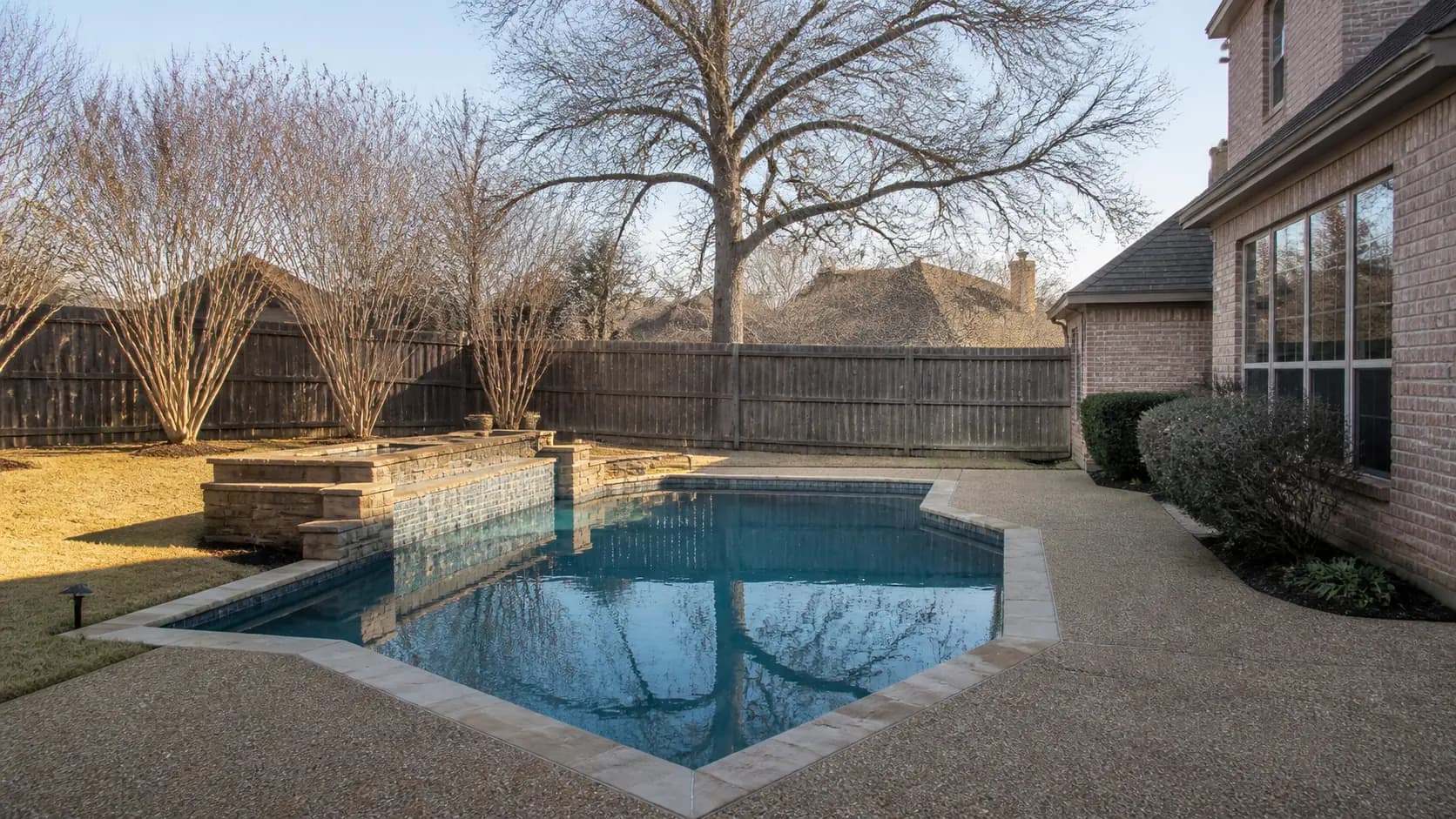 A clear suburban North Texas backyard pool under a low January sun, with bare crepe myrtles, dormant straw-colored grass, and a brick house wall behind.